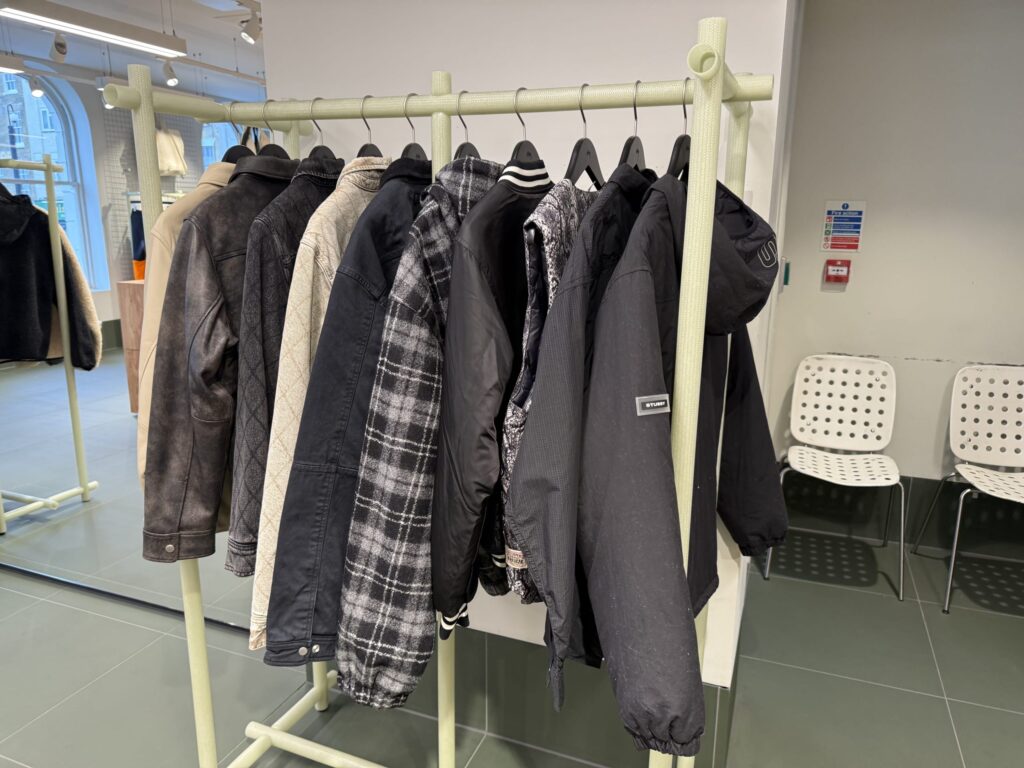 A variety of outerwear, including leather and plaid jackets, hanging on a light green clothing rack inside a select shop.