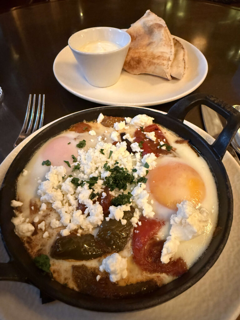 Shakshuka in a black cast iron skillet. It features two sunny-side-up eggs on a bed of tomato and pepper sauce, topped with crumbled feta cheese and parsley. A plate with pita bread and a white dipping sauce is in the background.