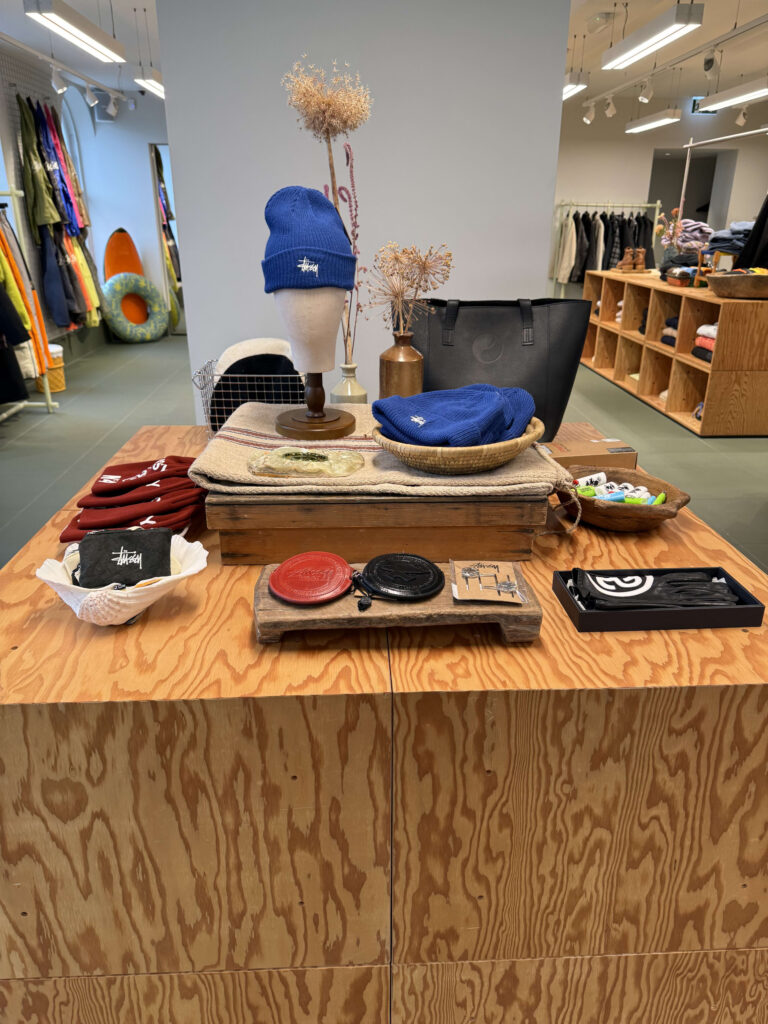 A view of fashion accessories such as a blue Stüssy beanie, a black tote bag, and leather coin purses displayed on a wooden table in a select shop.