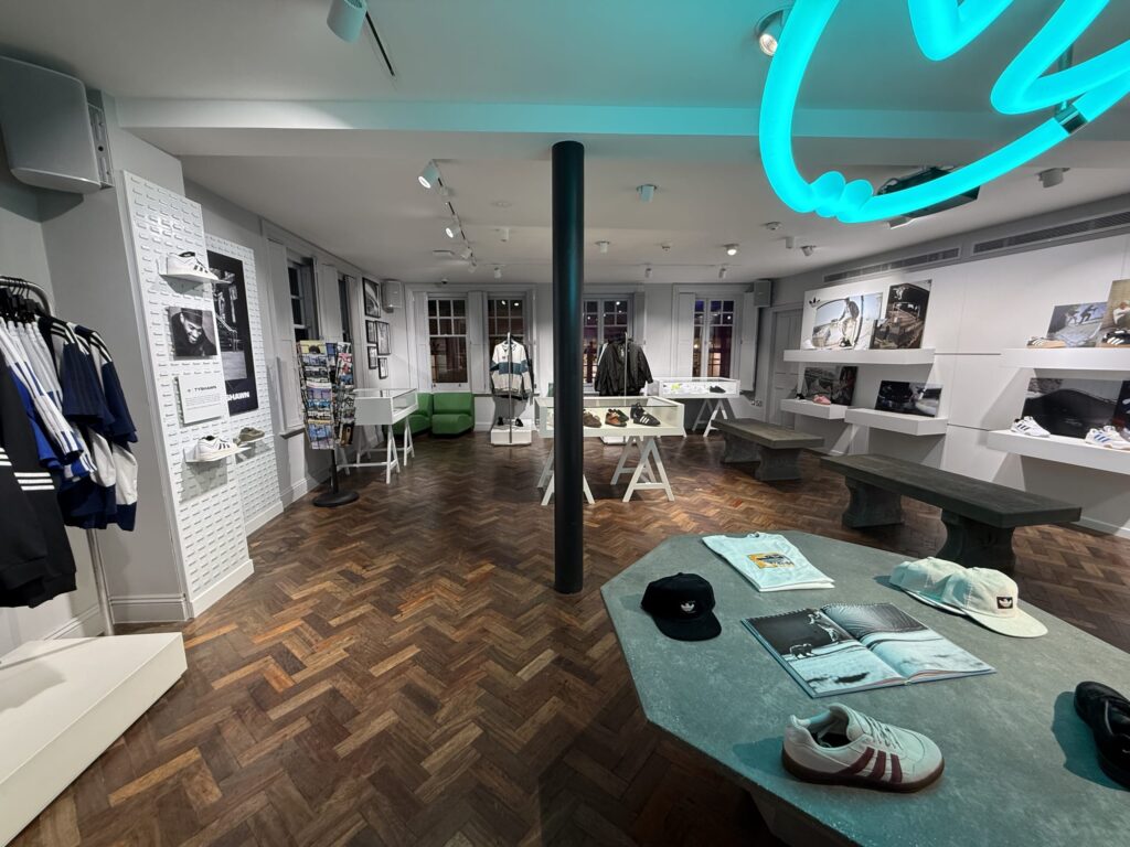 The interior of an Adidas store's exhibition space. Inside the store with herringbone patterned floors, apparel and sneakers from the skateboard collection are displayed, and a turquoise neon sign from the ceiling illuminates the space.
