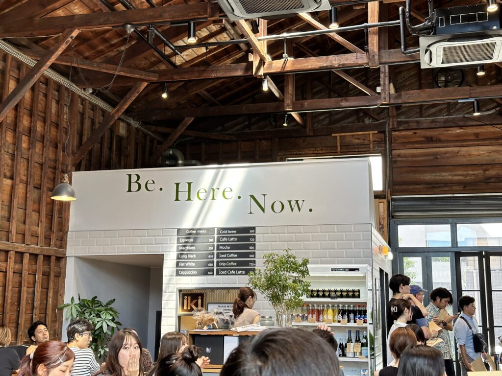The interior of a bustling cafe with a high ceiling and exposed wooden beams. A white counter has a sign above it that reads, 'Be. Here. Now.' and many customers are gathered around it.
