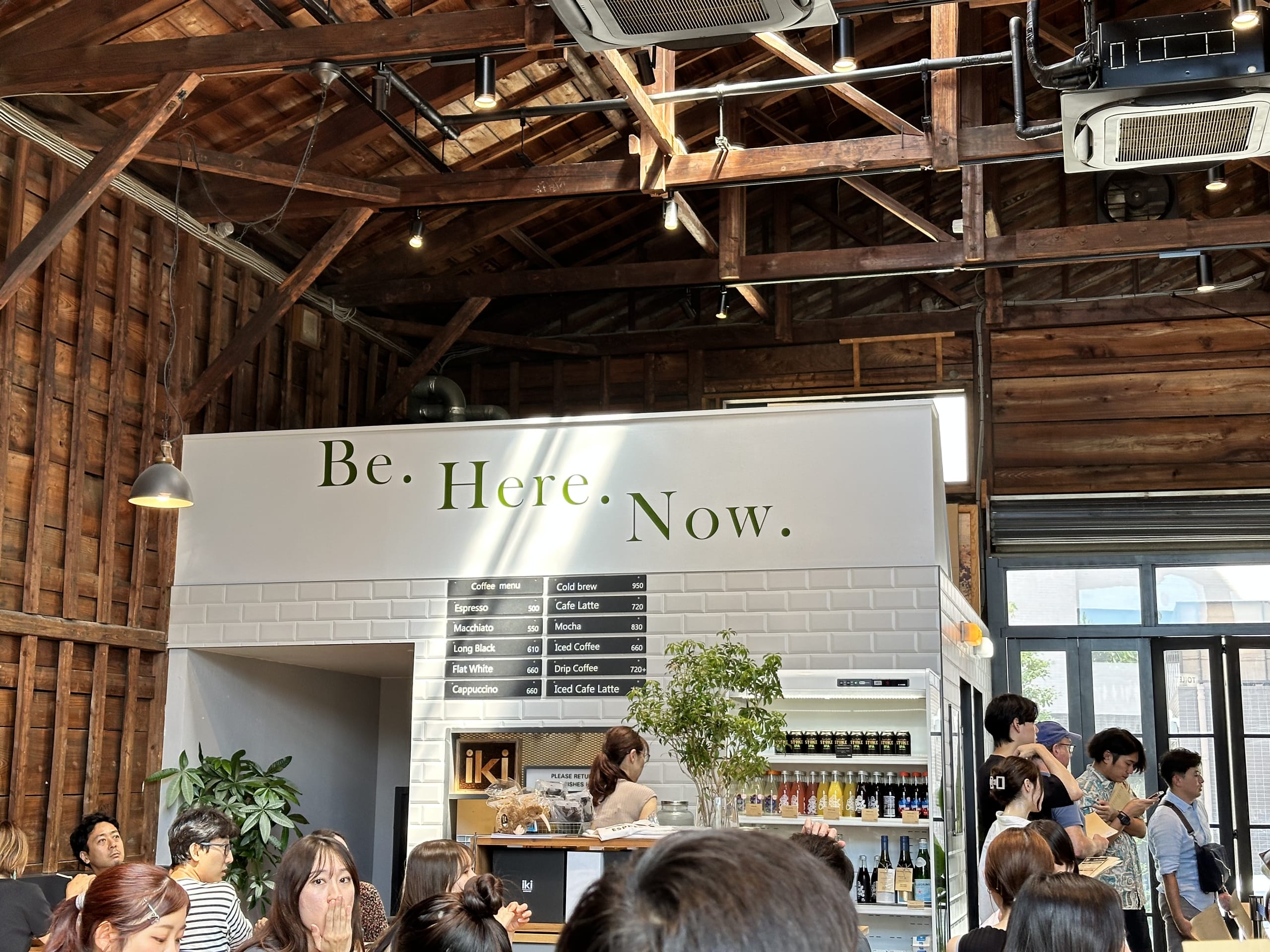 木の梁が印象的な高い天井のカフェ。白いカウンターの上には「Be. Here. Now.」と書かれた看板があり、大勢の客で賑わっている。