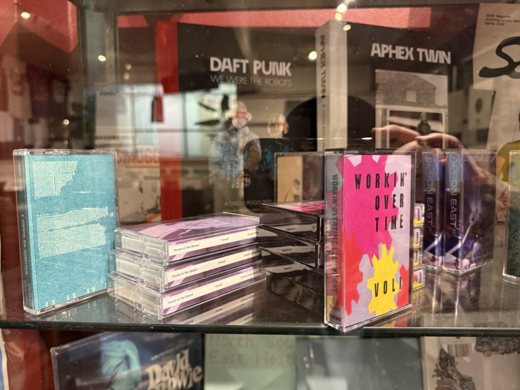 Cassette tapes displayed in a glass case at a record store. In the background, books and records by Daft Punk and Aphex Twin are visible.