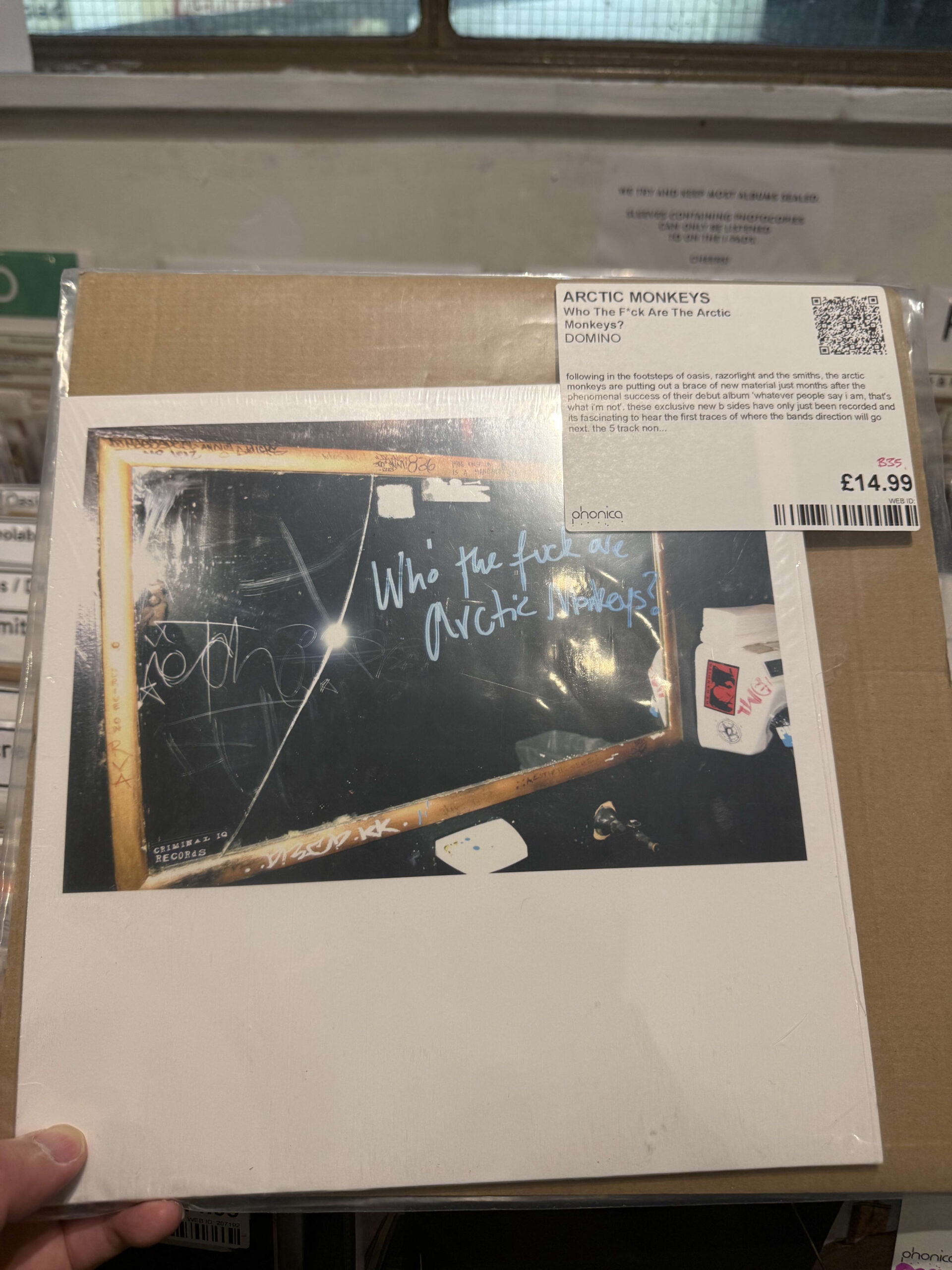 レコード店で手に持たれているアークティック・モンキーズのEPレコード「Who the F**k Are Arctic Monkeys?」。ジャケットには黒板にチョークで書かれたようなタイトルが写っている。