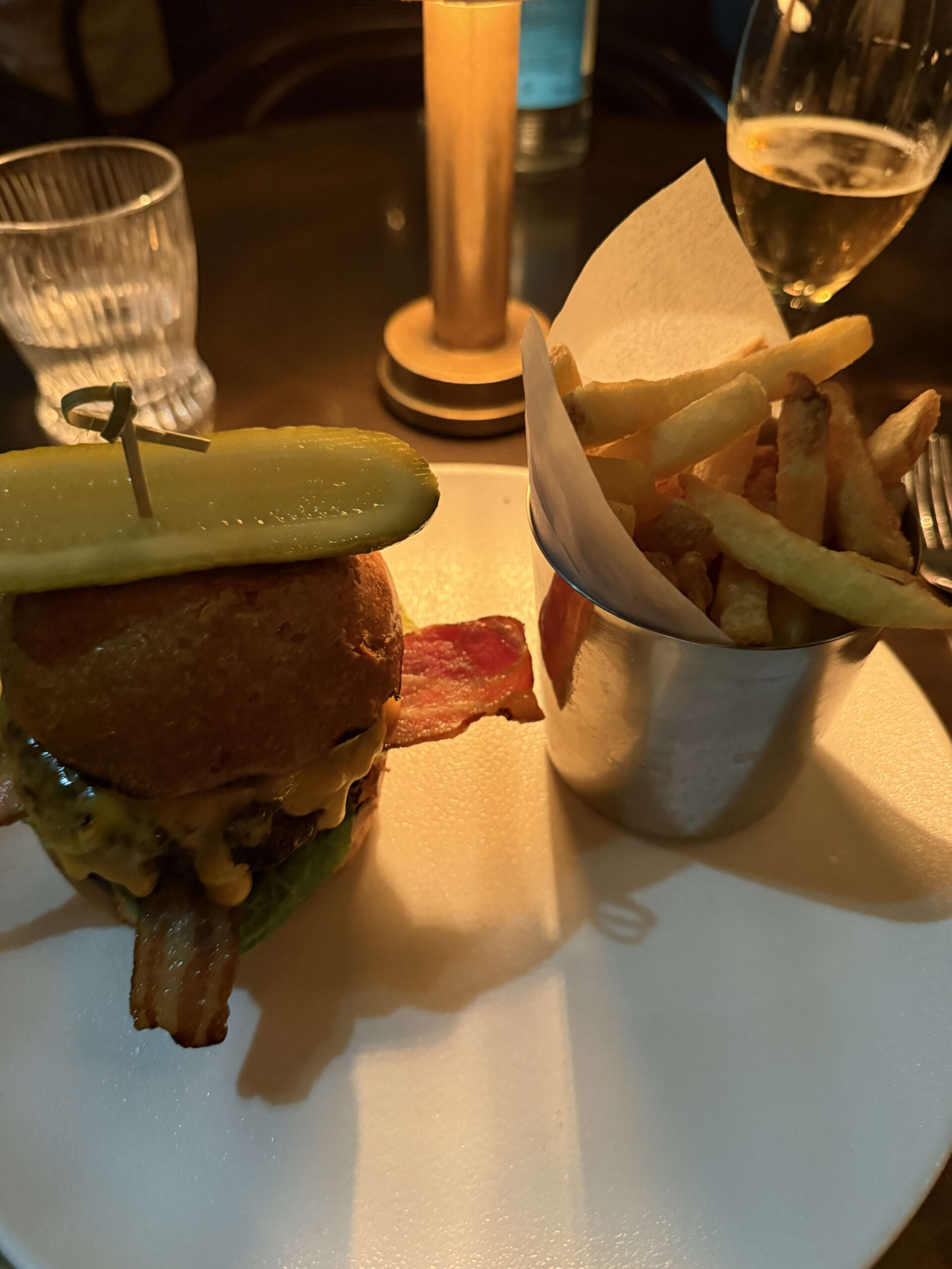 大きなピクルスが乗ったベーコン入りスマッシュバーガーとカップに入ったフライドポテト