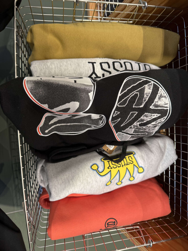 A view of folded graphic hoodies in various colors like yellow, grey, black, and red, stacked inside a wire basket.