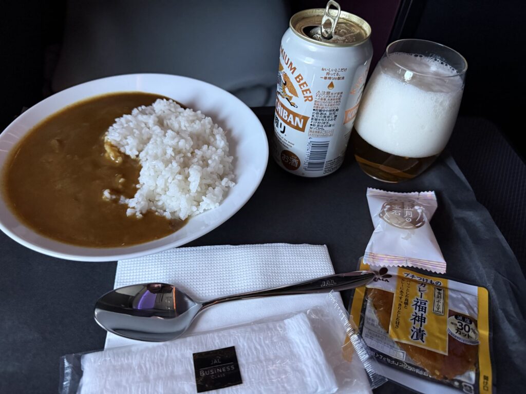JALビジネスクラスの機内食。白い皿に盛られたカレーライス、キリン一番搾りの缶ビールとグラス、福神漬け、デザートがトレーに並んでいる。