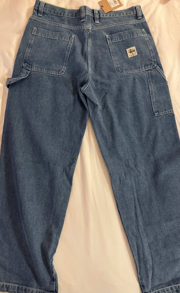 The back of a pair of Stüssy blue denim painter pants laid flat on a white surface, showing the back pockets, hammer loop, and a Stüssy workwear logo patch on the right pocket.