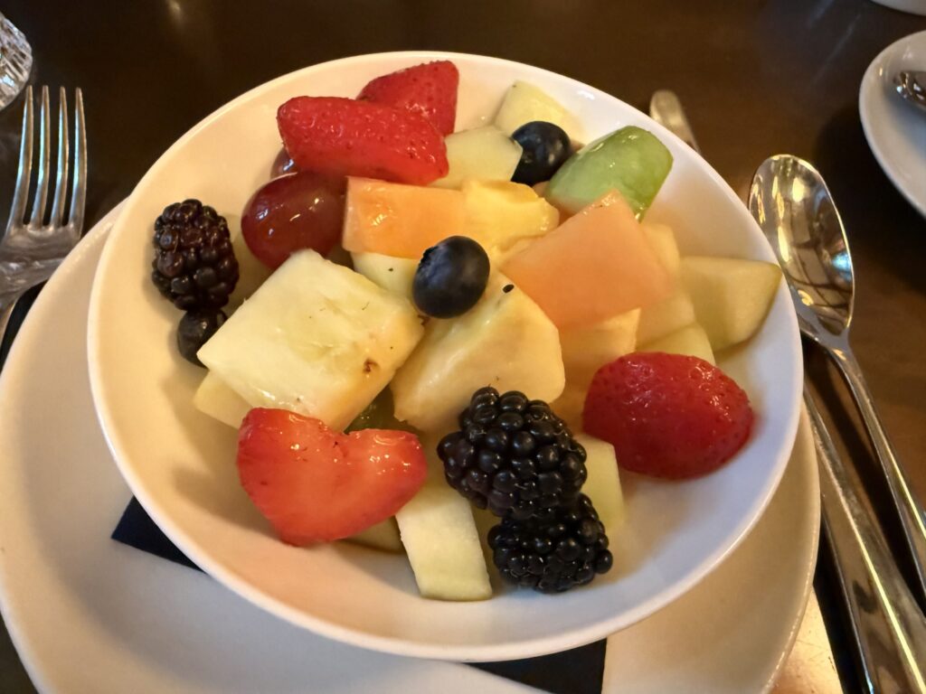 A white bowl filled with a colorful variety of cut fruits, including strawberries, blueberries, blackberries, pineapple, and melon. It is placed on a table next to a fork and spoon.