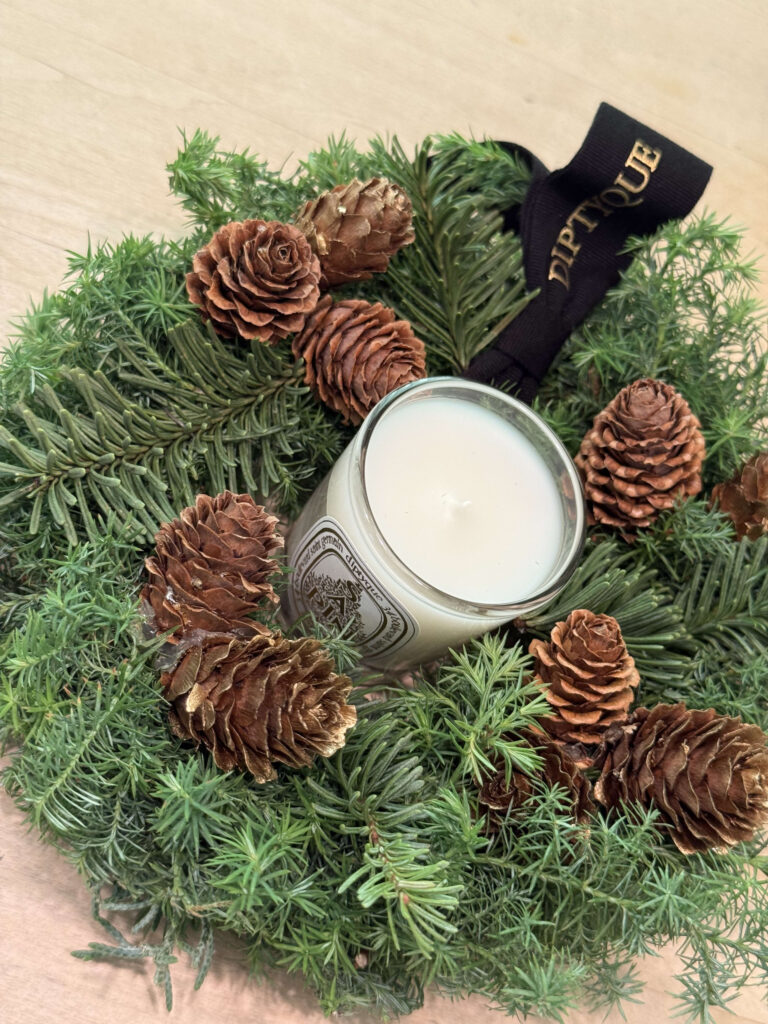 A Diptyque candle placed in the center of a real Christmas wreath decorated with pinecones on a wooden surface.