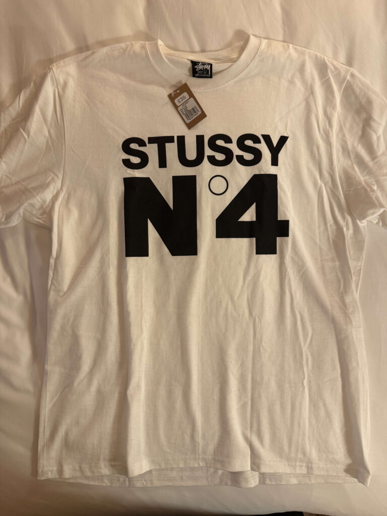 平置きされた白いStüssyのTシャツ。胸の中央に黒い太字で「STUSSY N°4」というロゴがプリントされている。首元には値段のタグが付いている。