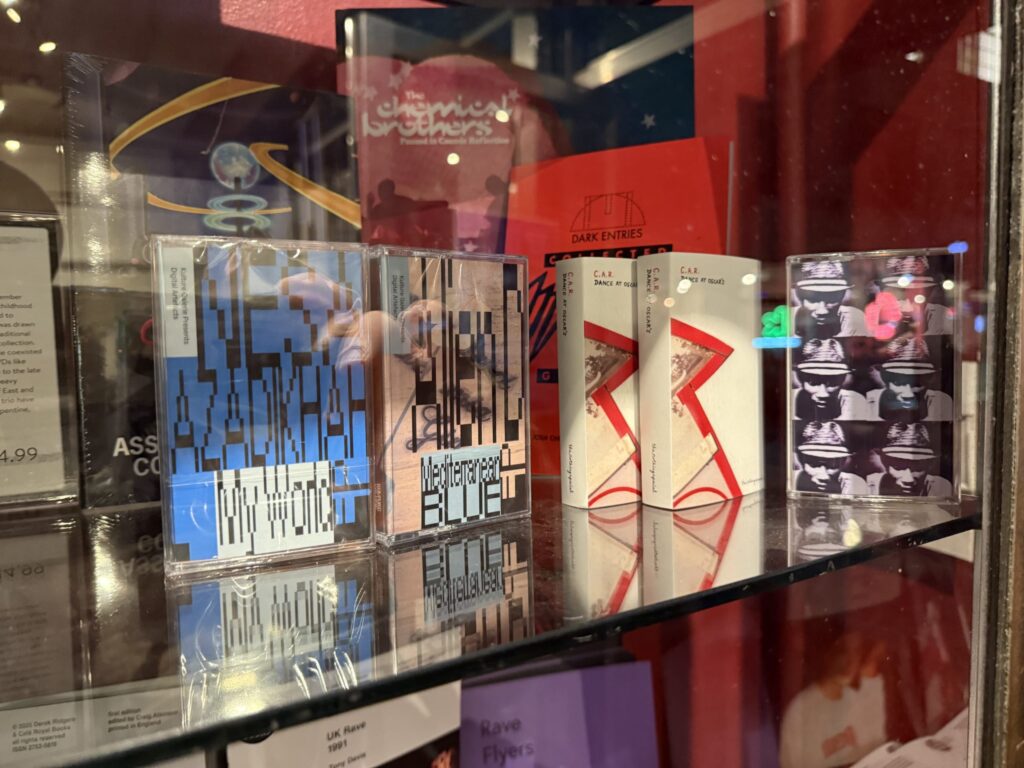 Graphically designed cassette tapes displayed on a glass shelf in a record store, with a Chemical Brothers CD visible in the background.