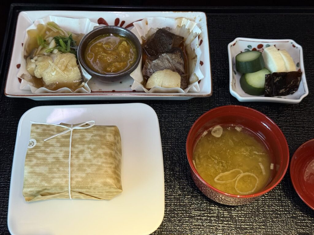 黒いお盆の上に並べられた和食のセット。主菜の皿には魚料理、肉料理、小鉢に入ったソースが乗っており、笹の葉で包まれたご飯、お味噌汁、香の物も添えられている。