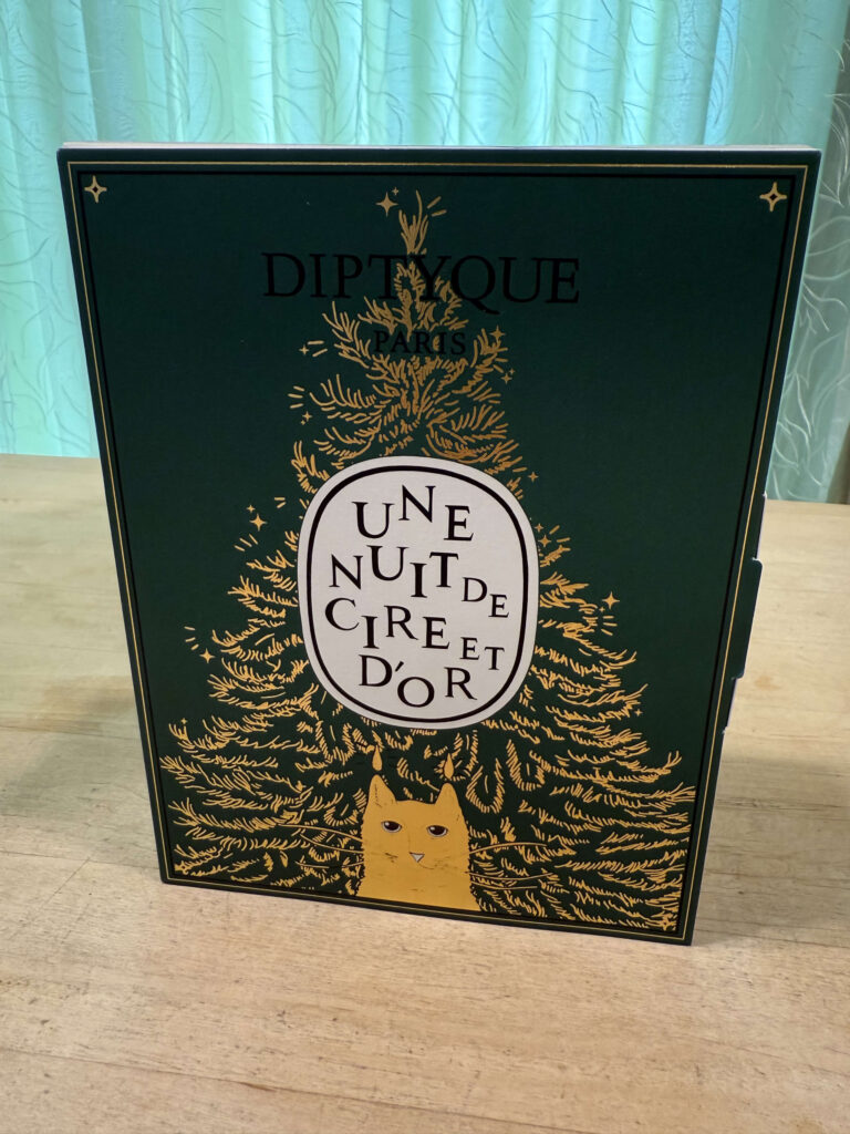 A Diptyque holiday collection box with a dark green background, featuring a gold Christmas tree and a yellow cat peeking out from the bottom.