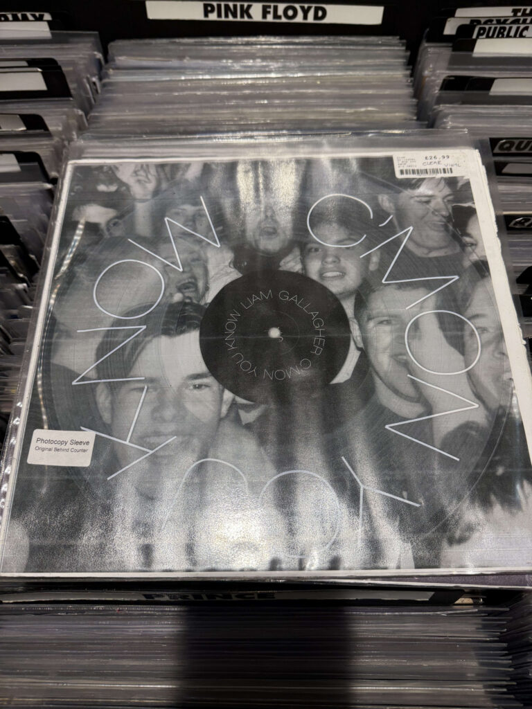 Liam Gallagher's 'C'MON YOU KNOW' record in a plastic sleeve, displayed in a record store. The cover is a monochrome collage of people's faces.