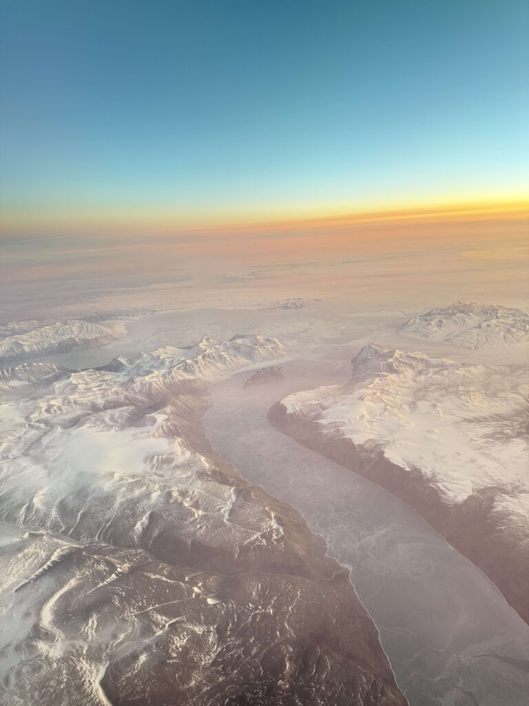 北極圏上空から撮影した、雪に覆われた山々と凍った川の空撮写真。地平線は夕焼けでオレンジ色に染まっている。