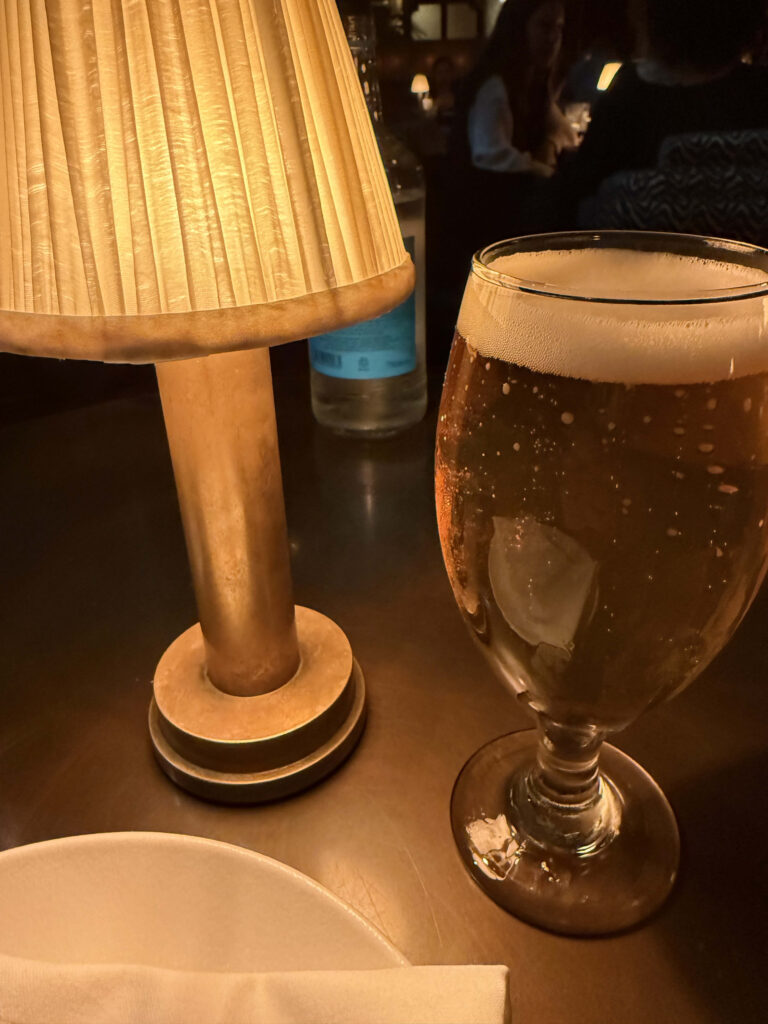 A glass of beer next to a warm table lamp in a dimly lit restaurant