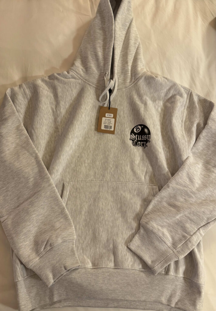 An ash heather Stüssy 8 Ball Corp. hoodie laid flat on a cream-colored background. The hoodie features a black 8-ball logo on the left chest.