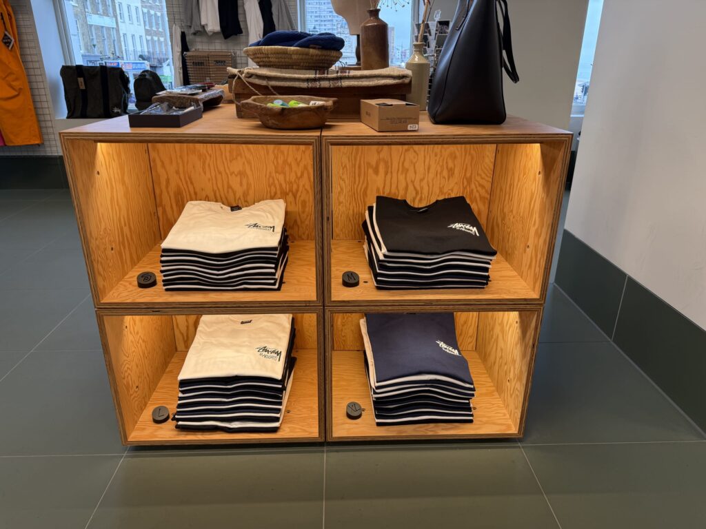 Stussy Margate店の店内。ライトアップされた木製の陳列棚の4つの区画に、白、黒、紺色のロゴ入りTシャツが綺麗に畳まれて陳列されている。
