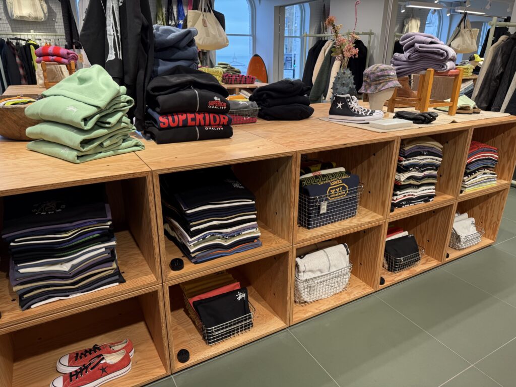 The interior of a clothing store, with neatly folded mint green and black sweatshirts, and various colored T-shirts arranged on a plywood display unit.