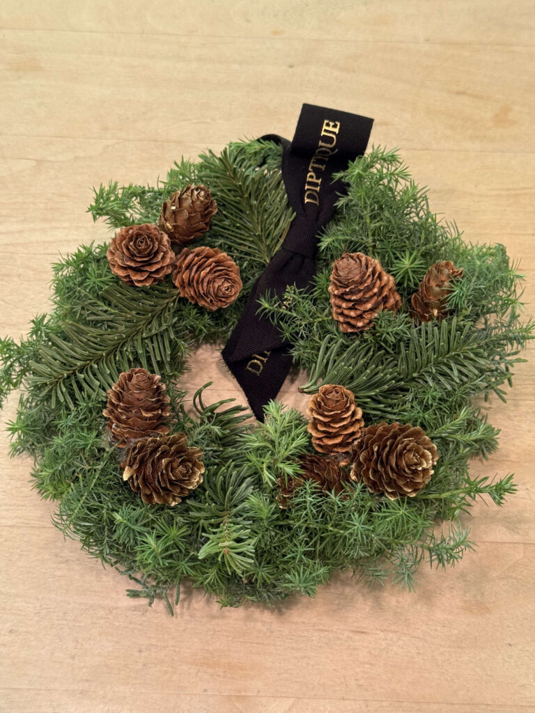 A Christmas wreath decorated with pinecones and a black Diptyque ribbon, placed on a wooden table.