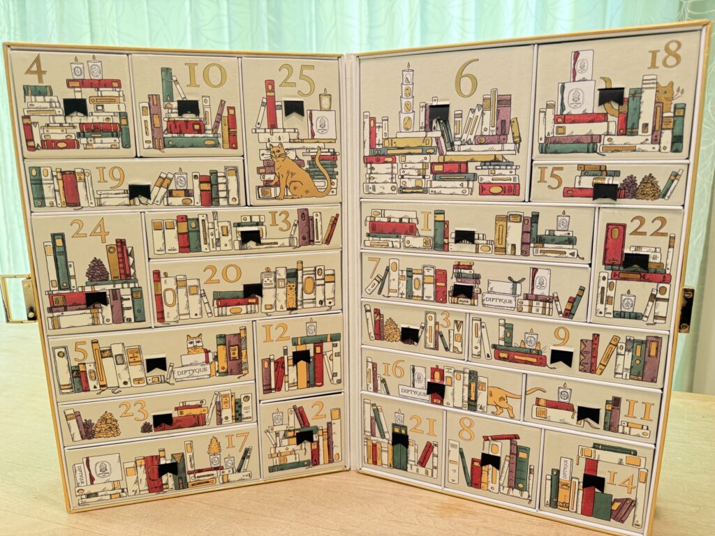 An open Diptyque advent calendar designed to look like a bookshelf, with 25 numbered drawers featuring illustrations of books, cats, and candles.