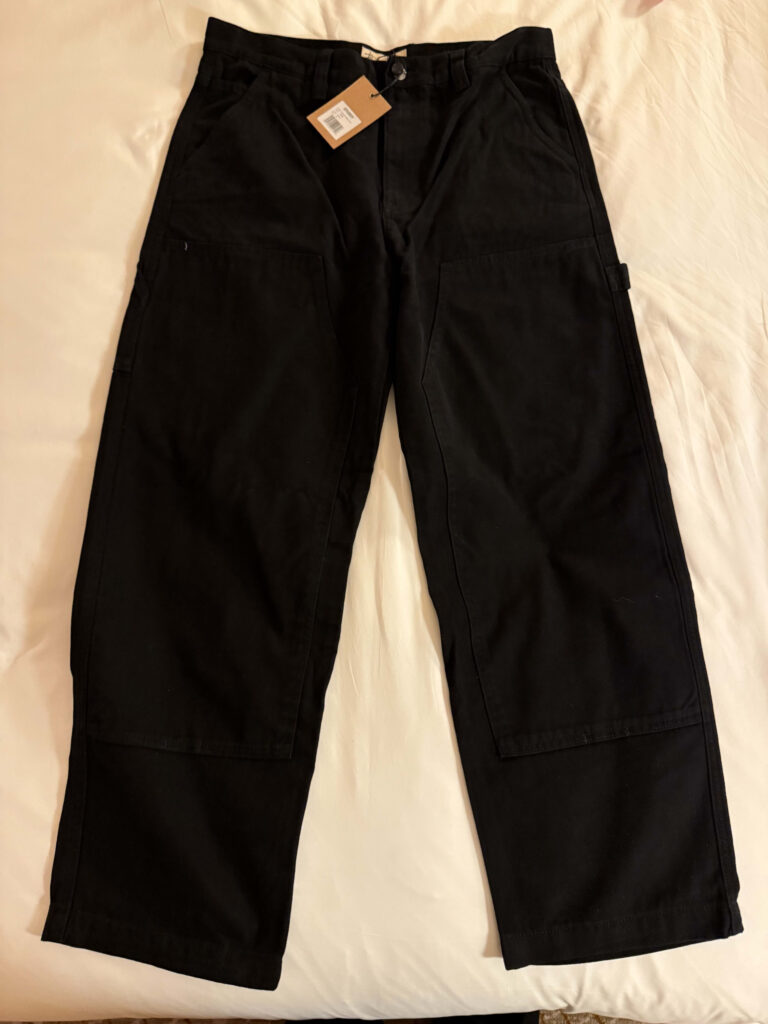 A pair of new, black Stüssy double knee work pants with tags still attached, laid flat on a white cloth surface.