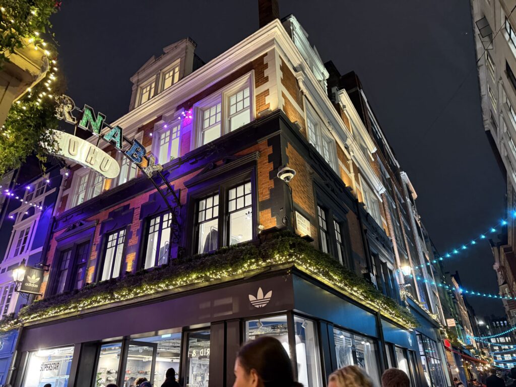 A low-angle shot of the exterior of the Adidas Originals flagship store on Carnaby Street in London at night. The brick building is illuminated with festive lights and purple uplighting.