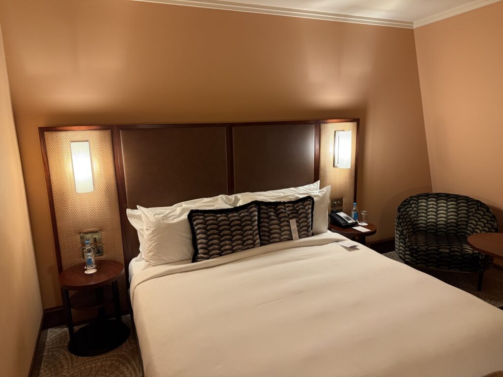 温かみのある照明に照らされたホテルの客室。中央にキングサイズベッドが置かれ、両脇にサイドテーブル、隅には模様の入ったアームチェアがある。