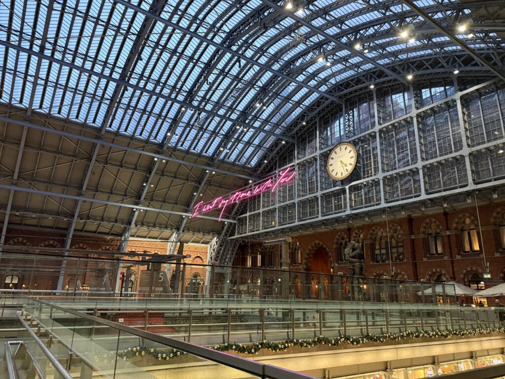 The interior of a grand train station with an arched glass ceiling. A pink neon sign that reads 'I want my time with you' hangs below the roof next to a large, traditional clock.