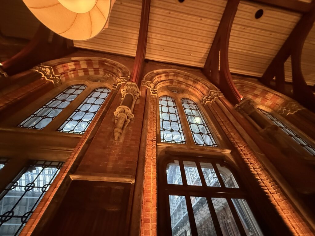 The interior of a historic building, illuminated by warm light. A low-angle shot looks up at red brick walls with large arched windows, revealing the steel roof of a station beyond.