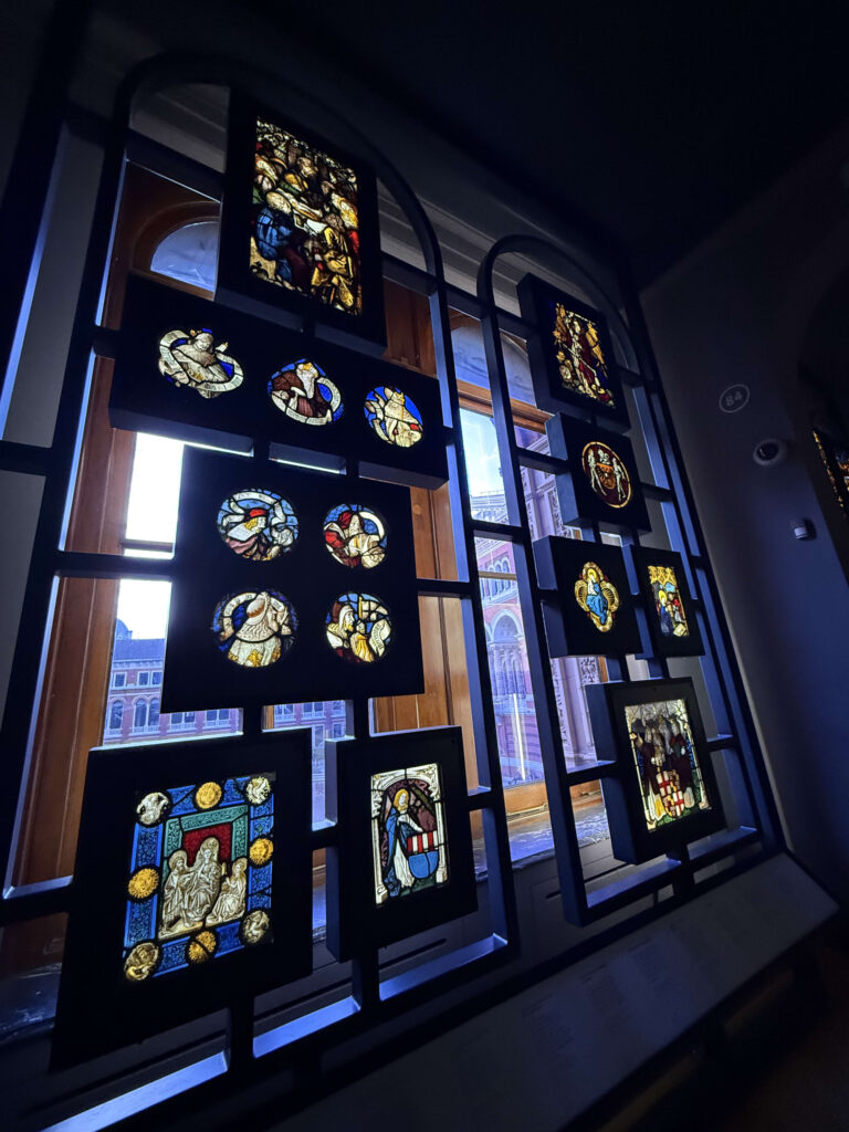 A collection of various stained glass panels of different shapes and designs displayed in black frames in front of a large window inside a dimly lit museum. The light from the window shines through the stained glass, making the colors glow vibrantly.