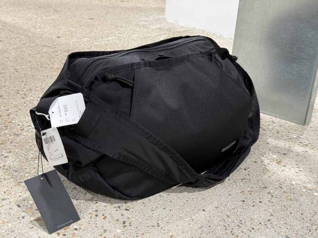 Black COMME des GARCONS HOMME x BRIEFING collaboration bag with tags sitting on a concrete floor