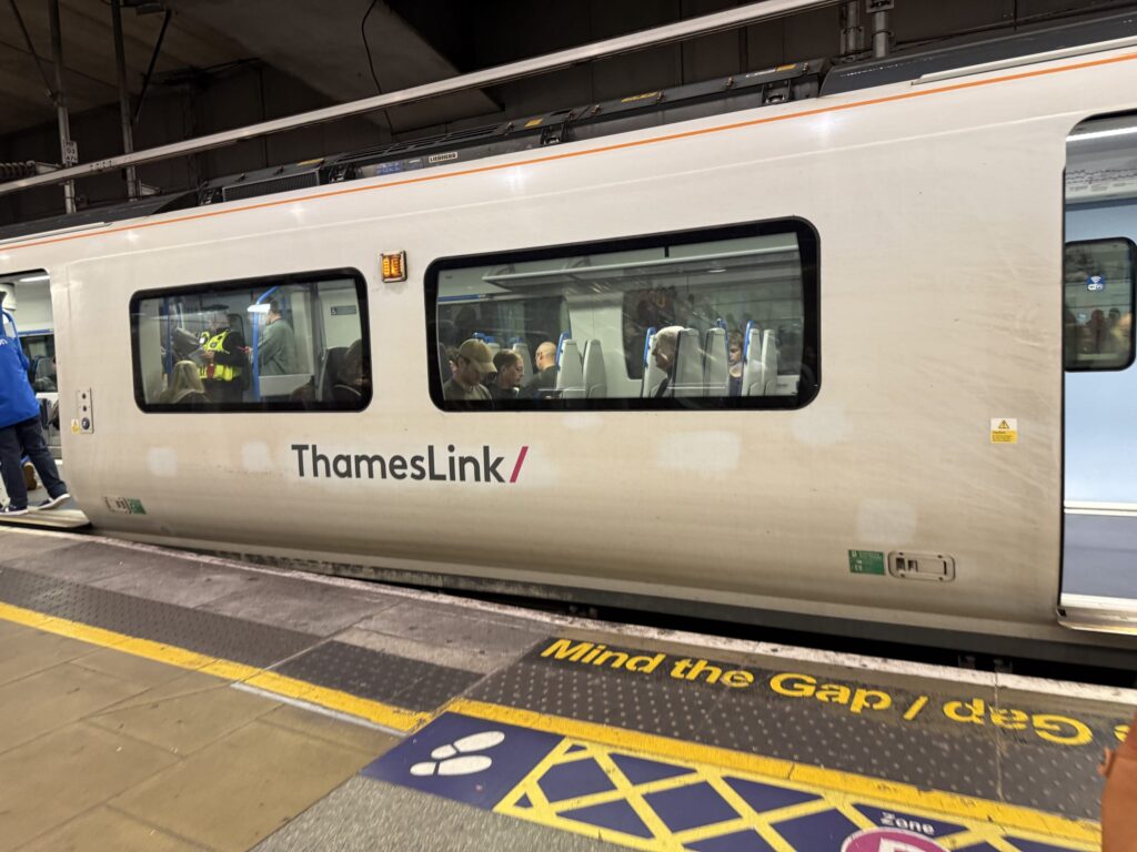 A white ThamesLink train stopped at a station platform with passengers visible through the windows and Mind the Gap text on the floor