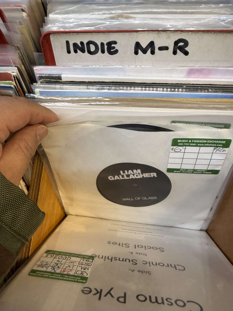 レコード店の「INDIE M-R」コーナーで、リアム・ギャラガーの『Wall of Glass』7インチシングルを手に取っている様子