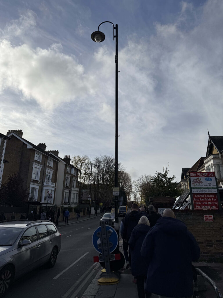 Street scene near Norwood Junction with a tall lamp post, people walking on the sidewalk, and brick houses under a cloudy sky.