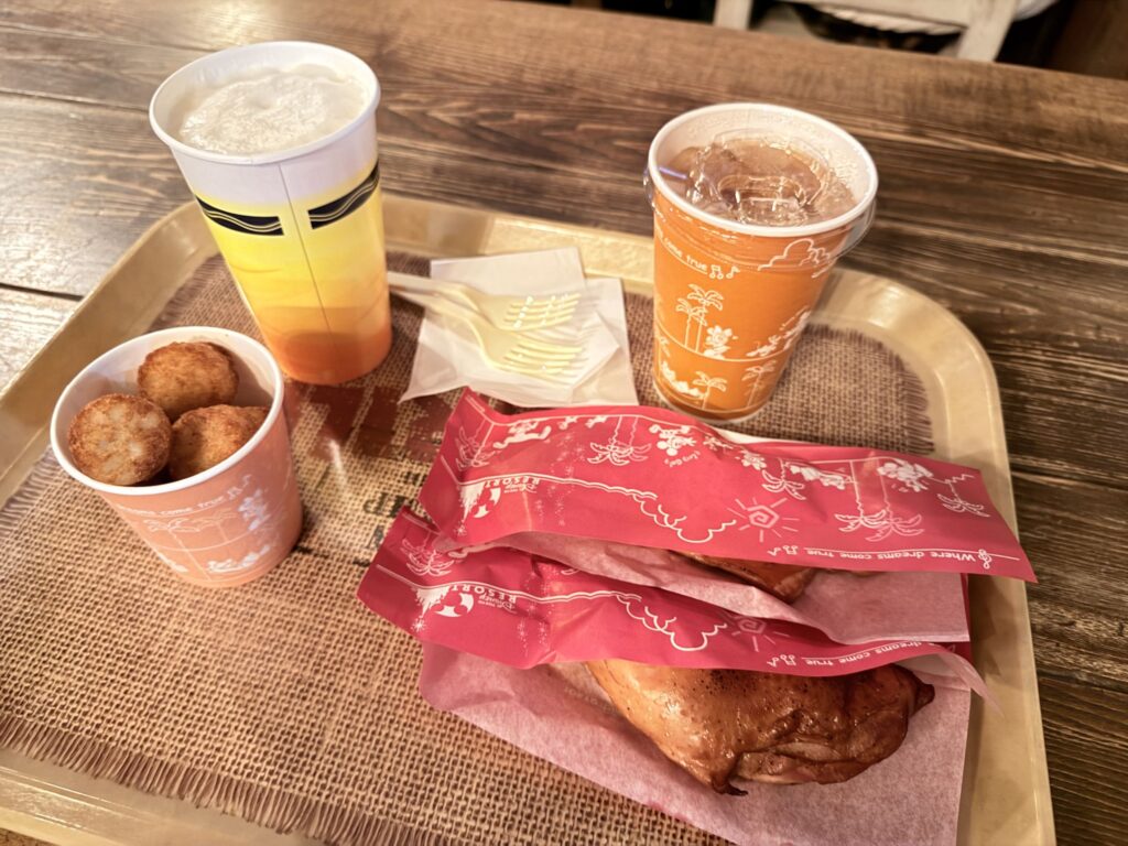 A food tray on a wooden table featuring spicy smoked chicken legs, onion hashed potatoes, a beer, and an apple tea soda