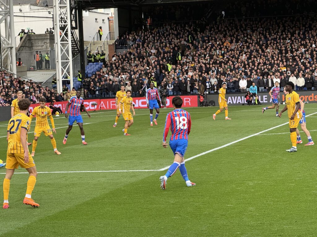 Rear view of Daichi Kamada wearing the number 18 Crystal Palace jersey on the pitch, facing Brighton players in yellow kits, with packed stadium stands in the background.