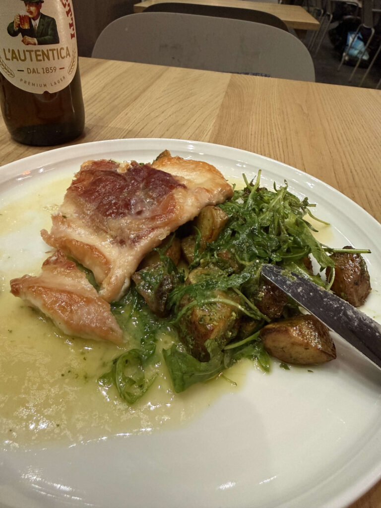 Grilled chicken wrapped in prosciutto served with roasted potatoes and arugula salad on a white plate, with a bottle of Moretti beer in the background