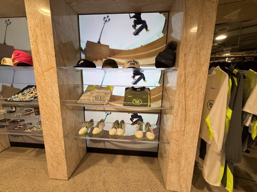 大理石調の棚に陳列されたNikeとPalaceのコラボレーション商品(キャップ、ジャージ、スニーカー)と、背景のスケートボードのビジュアル