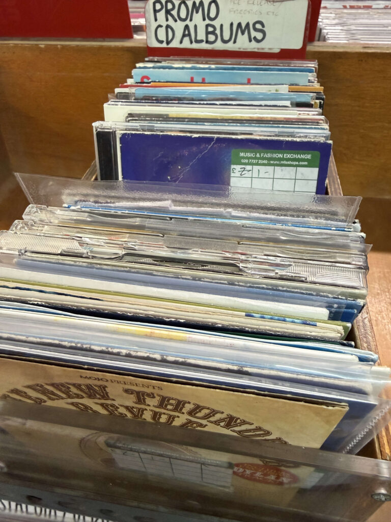 「PROMO CD ALBUMS」という手書きのサインが掲げられた木製の棚に、プロモーション用や雑誌付録などのCDが多数並んでいる様子