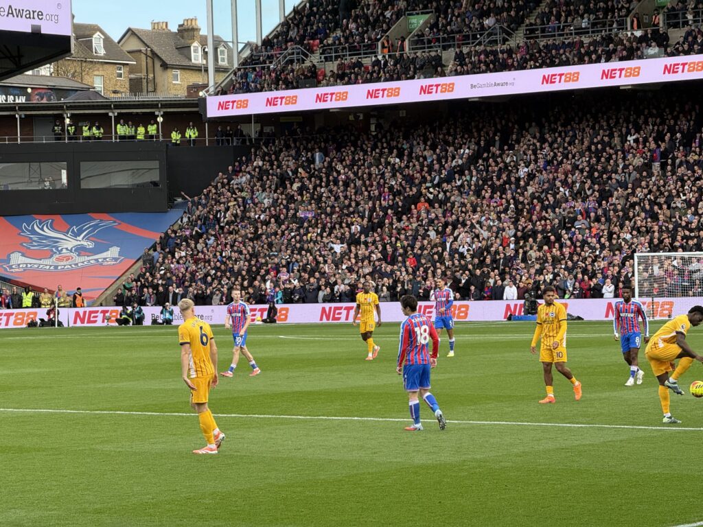 Crystal Palace player Daichi Kamada, wearing number 18, standing on the pitch at a crowded Selhurst Park stadium during a match against a team in yellow kits