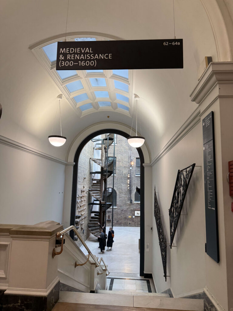美術館のホールから階段を見下ろした景色。頭上には「MEDIEVAL & RENAISSANCE」の案内板があり、アーチ型の通路の先にはレンガの壁に囲まれた中庭と、そこに設置された木製の螺旋階段が見える。