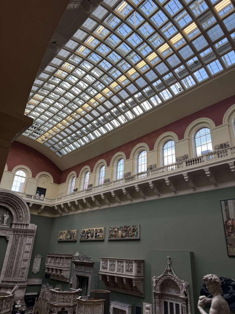 An upward view of a spacious museum gallery. Natural light streams in through an arched glass ceiling, illuminating the European sculpture and architectural casts lining the walls.