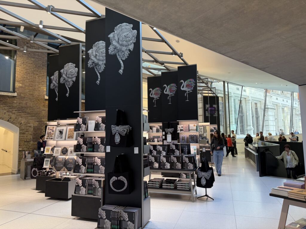 The interior of a special museum gift shop for a Cartier exhibition. Black display units and pillars are decorated with large graphics of jewelry, such as diamond roses and flamingos. Related books, tote bags, and other merchandise are on display.