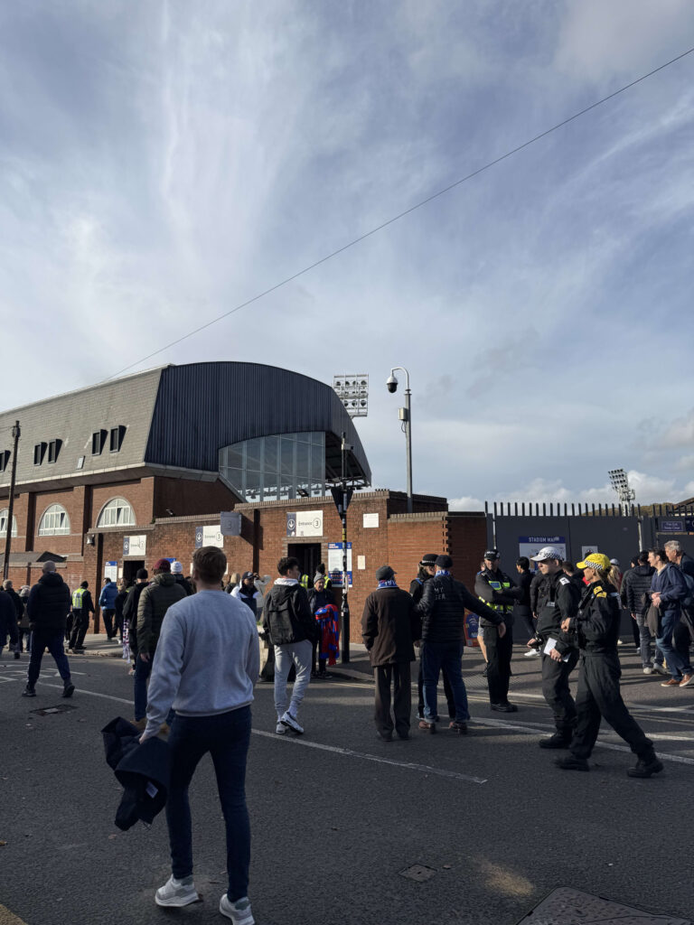 Crowd of fans walking towards a brick stadium entrance guarded by police officers under a blue sky
