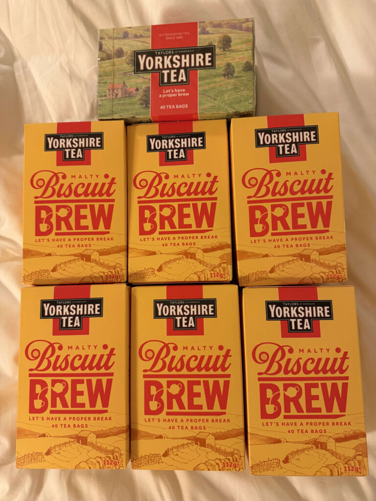 A collection of Taylors of Harrogate tea boxes arranged on a white sheet, featuring one red box of original Yorkshire Tea on top and six yellow boxes of Malty Biscuit Brew in two rows below.