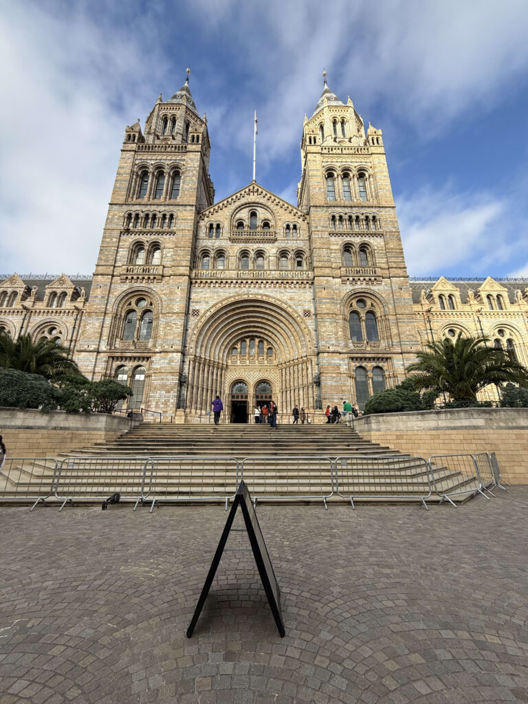 The grand front facade of the Natural History Museum in London under a blue sky, featuring two towers and a large arched entrance.