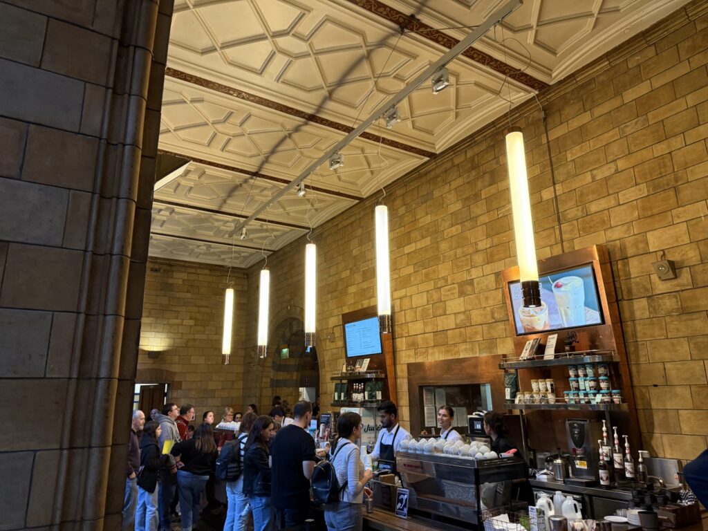 A busy cafe counter inside a historic brick building with high decorative ceilings and hanging modern tube lighting, showing a long line of customers waiting to order