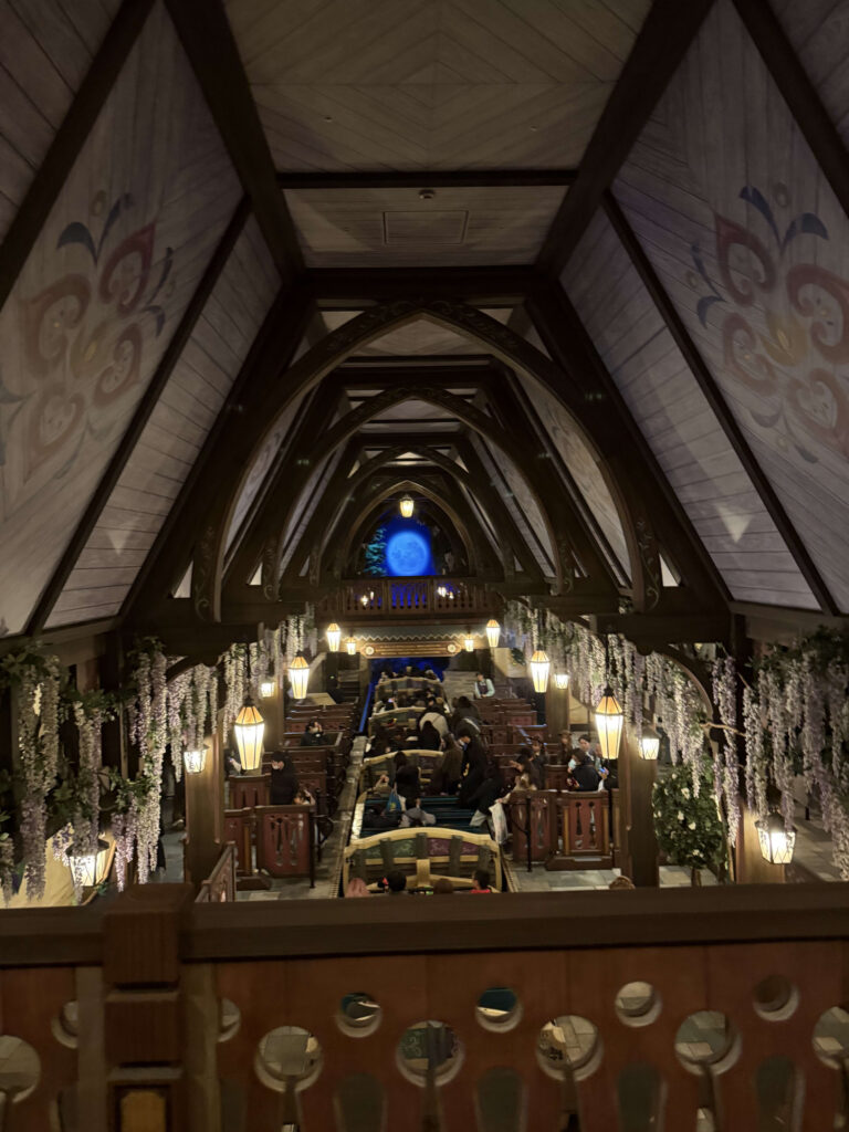 High-angle view of an indoor boat ride boarding area featuring wooden arches with painted designs, hanging wisteria flowers, and warm lantern lighting leading to a blue-lit tunnel.